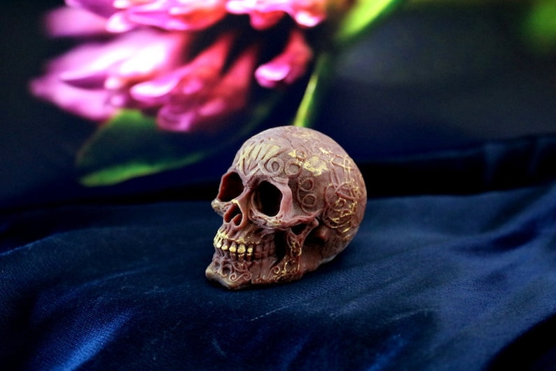 May include: A detailed, brown skull figurine with gold accents. The skull has intricate carvings and is set against a dark blue background. The teeth are a gold colour. The overall theme is gothic or macabre.