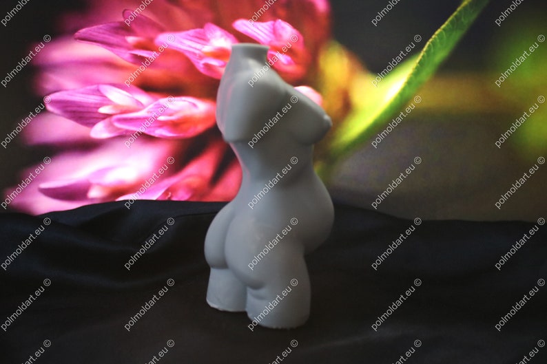 May include: A light grey, sculpted torso figurine. The form depicts a female figure with defined breasts and a rounded abdomen. The background features a blurred floral pattern in shades of pink and green, with the website address "polmoldart.eu" repeated.