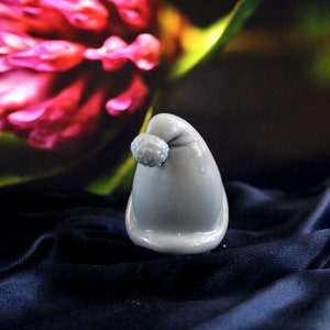 May include: A small, gray, Santa hat-shaped ornament with a pom-pom on top. The ornament is glossy and sits on a dark blue, textured fabric. A blurred background features pink and green floral elements.