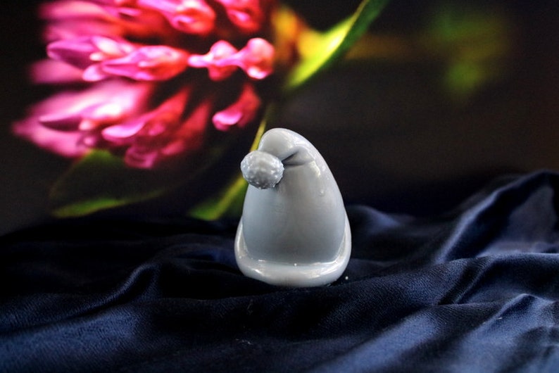 May include: A small, gray, Santa hat-shaped ornament with a pom-pom on top. The ornament is glossy and sits on a dark blue, textured fabric. A blurred background features pink and green floral elements.