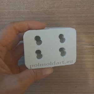 Puede incluir: Un molde rectangular blanco con cuatro agujeros circulares. El texto "polmoldart.eu" est&aacute; impreso en el borde inferior. El molde se sostiene en una mano.