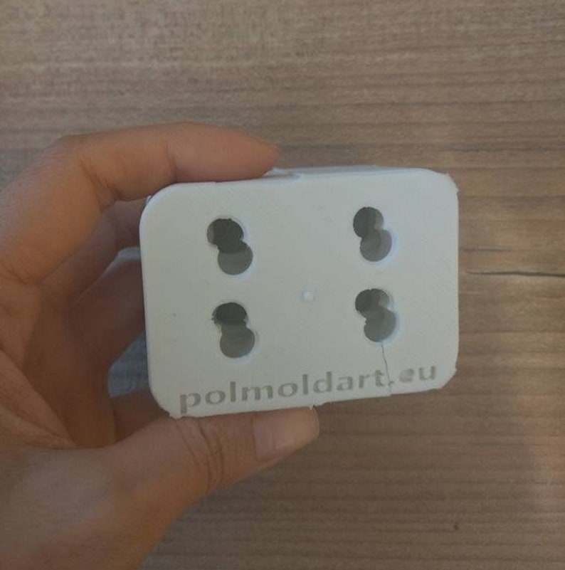 Puede incluir: Un molde rectangular blanco con cuatro agujeros circulares. El texto "polmoldart.eu" est&aacute; impreso en el borde inferior. El molde se sostiene en una mano.