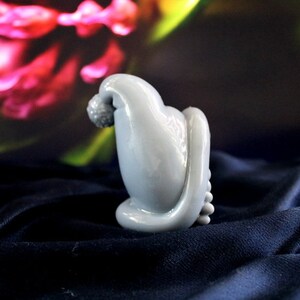 May include: A light blue, abstract sculpture with a rounded top and a textured, spherical element. The sculpture has a smooth, glossy finish and sits on a dark blue, textured surface. The background features blurred pink and green floral elements.