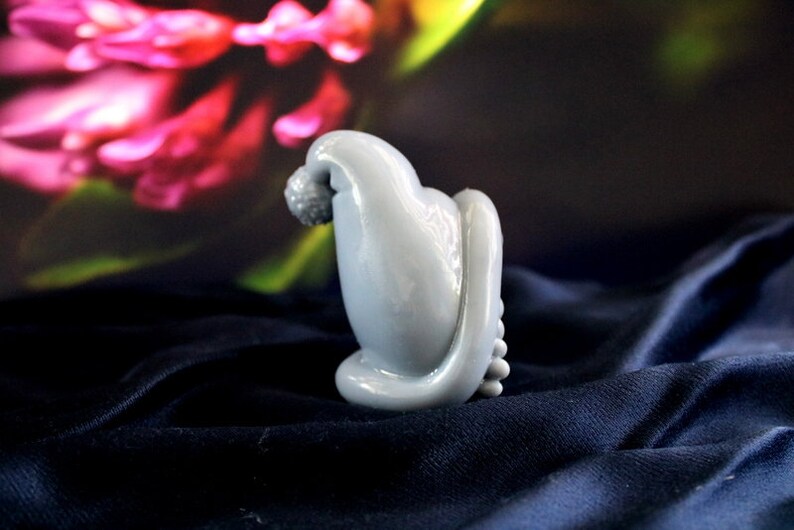 May include: A light blue, abstract sculpture with a rounded top and a textured, spherical element. The sculpture has a smooth, glossy finish and sits on a dark blue, textured surface. The background features blurred pink and green floral elements.