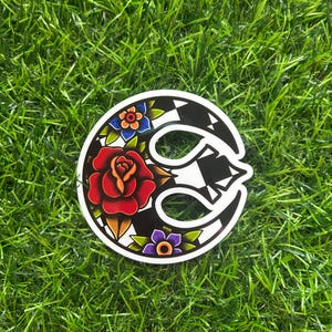 May include: A circular sticker with a black and white design and floral accents. A red rose is in the center, with blue and purple flowers around the edge. The sticker is on a bed of green grass.