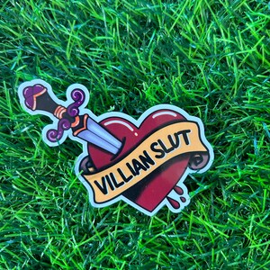May include: A sticker featuring a heart pierced by a sword. The heart is burgundy with a banner that reads "VILLIAN SLUT". The sword has a purple handle and a silver blade. The sticker has a white border.