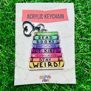 May include: Acrylic keychain featuring a stack of colorful books with the phrases "Read Books", "Be Kind", and "Stay Weird!". The keychain has a silver-colored clasp and is displayed on a brown card.