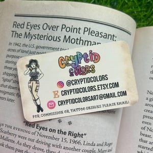 May include: A rectangular business card with rounded corners featuring the colorful text Cryptid Colors, an illustration of a person in a dark top and light pants, and contact information including an Instagram handle, Etsy URL, and email address.