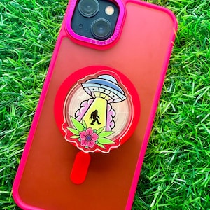 May include: A bright red smartphone case with a matching red circular phone grip attached to the back. The grip features a graphic illustration of a UFO beaming down a silhouette of Bigfoot, surrounded by pink clouds and a green floral design.
