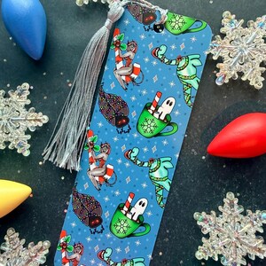 May include: A rectangular bookmark with a blue background featuring a repeating pattern of holiday-themed illustrations including ghosts in green mugs, figures on candy canes, and moths wrapped in string lights, finished with a silver tassel.