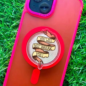 May include: A bright pink smartphone case with a circular phone grip attached to the back. The grip features a graphic of a tan ribbon with the text "YOU WILL NOT BREAK ME!" printed in black capital letters across the center of the ribbon.