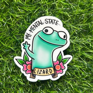 May include: A sticker featuring a cartoon lizard with the text "MY MENTAL STATE" and "LIZARD". The lizard is green with large eyes and is giving a thumbs up. Pink flowers and green leaves surround the lizard.