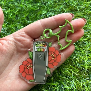 May include: A clear acrylic keychain featuring a gray ghost meter with a red, yellow, green, and blue gauge. The gauge reads "milligauss" and has an arrow pointing to the "high" reading. The keychain also features two red flowers with yellow centers.