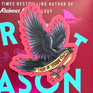 May include: A die-cut sticker featuring a black crow with spread wings, set against a vibrant pink background. The crow holds a yellow banner in its talons with the black text TASTE OF TERROR FESTIVAL printed clearly across the ribbon surface.