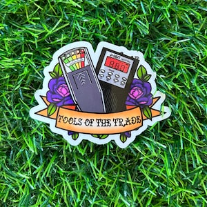 May include: A sticker with a cartoon illustration of a ghost hunting kit. The kit includes a P-SB7 Spirit Box and a EMF meter. The sticker has a banner that reads "Tools of the Trade".