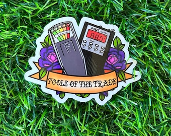 Puede incluir: Una pegatina con una ilustración de dibujos animados de un kit de caza de fantasmas. El kit incluye una caja de espíritus P-SB7 y un medidor EMF. La pegatina tiene una pancarta que dice "Tools of the Trade".