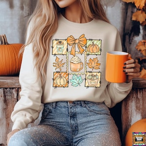 May include: Cream-colored sweatshirt featuring a grid of fall-themed illustrations, including pumpkins, leaves, and coffee. An orange bow is centered at the top. The person is holding an orange mug. The sweatshirt is paired with blue jeans.