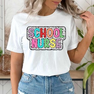 May include: White t-shirt with a colorful "School Nurse" graphic. The graphic has a black and white polka dot background.