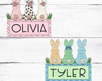 May include: Three Easter bunny-shaped decorations with different patterns and colors. The bunnies are sitting on a green grass patch with a pink and white sign that says "Olivia". Below them are three more bunnies with different patterns and colors sitting on a green grass patch with a blue and green sign that says "Tyler".