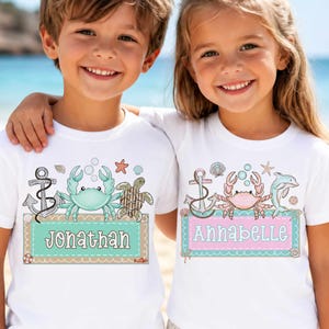 May include: Two white t-shirts with matching sea-themed graphics. One shirt features a teal crab, the name "Jonathan" on a teal banner, and other sea elements. The other shirt has a pink crab, the name "Annabelle" on a pink banner, and a dolphin.