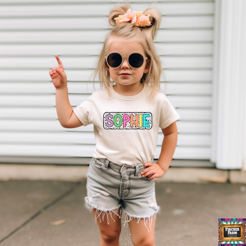 May include: A young girl wearing a white t-shirt with a colorful "SOPHIE" graphic on a black and white polka dot background. She is wearing denim shorts and sunglasses.