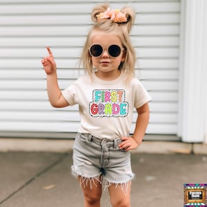 May include: A young girl wearing a white t-shirt with the words "FIRST GRADE" in rainbow colors on a black and white polka dot background. She is wearing denim shorts and sunglasses.