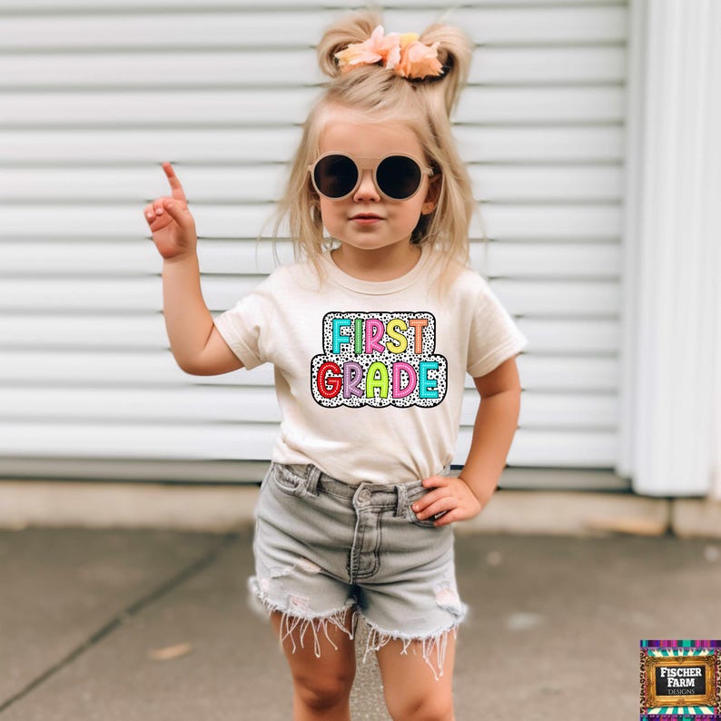 May include: A young girl wearing a white t-shirt with the words "FIRST GRADE" in rainbow colors on a black and white polka dot background. She is wearing denim shorts and sunglasses.