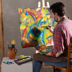 Pu&ograve; includere: Una persona dipinge su una tela con un'opera d'arte astratta e colorata. La tela &egrave; su un cavalletto. La persona tiene un pennello nella mano destra. Ci sono tubetti di vernice e pennelli in un vaso di terracotta marrone su un tavolo. C'&egrave; una tavolozza di legno con un coltello da pittura sopra.