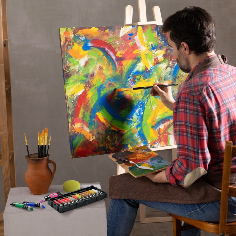 Pu&ograve; includere: Una persona dipinge su una tela con un'opera d'arte astratta e colorata. La tela &egrave; su un cavalletto. La persona tiene un pennello nella mano destra. Ci sono tubetti di vernice e pennelli in un vaso di terracotta marrone su un tavolo. C'&egrave; una tavolozza di legno con un coltello da pittura sopra.