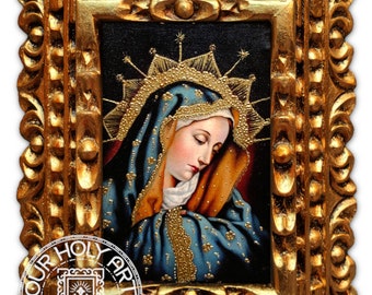 May include: A framed religious icon featuring a painting of a woman in a blue and gold head covering. The painting is set within an ornate, gold-colored frame. The frame has a detailed, textured design. The image includes the text "OUR HOLY ART.COM".