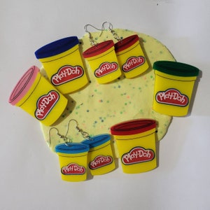 May include: Six pairs of Play-Doh themed earrings. Each pair features a small plastic container shaped like a Play-Doh container with the Play-Doh logo. The containers are in various colors, including yellow, red, blue, and green.