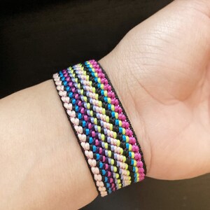 May include: A colourful woven bracelet with pink, blue, green, and black stripes. The bracelet is made of a thin, braided material and is worn on a person's wrist.