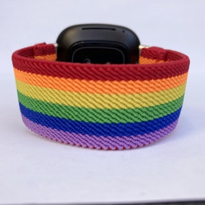 May include: A rainbow-striped watch band in red, orange, yellow, green, blue, and purple. The band is made of a woven material and is wrapped around a black smartwatch. The band is designed to fit a smartwatch.