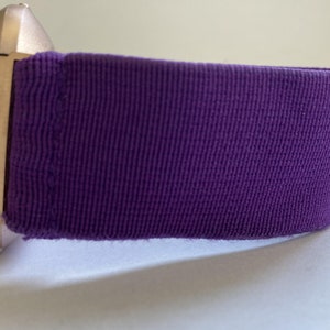 Pu&ograve; includere: Primo piano di uno smartwatch con cinturino in tessuto viola. La cassa dell'orologio &egrave; color oro chiaro. Il cinturino &egrave; di un viola intenso e sembra essere realizzato in un tessuto strutturato.