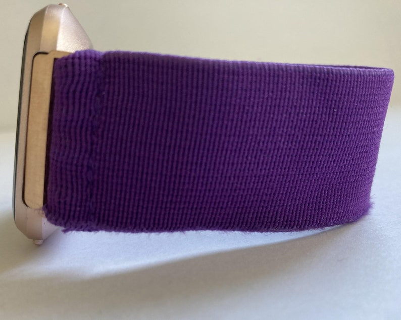 Pu&ograve; includere: Primo piano di uno smartwatch con cinturino in tessuto viola. La cassa dell'orologio &egrave; color oro chiaro. Il cinturino &egrave; di un viola intenso e sembra essere realizzato in un tessuto strutturato.