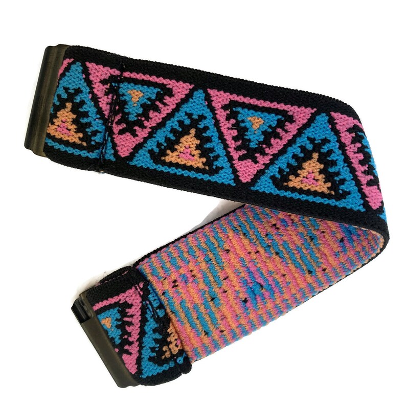 May include: A black elastic guitar strap with a colorful geometric pattern. The pattern features pink, blue, and orange triangles with black outlines.