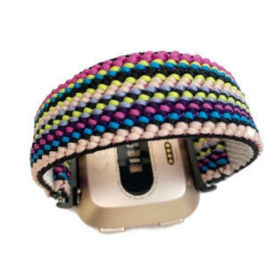 May include: A colourful woven watch band with pink, purple, blue, green, black and white stripes. The band is attached to a gold-coloured smartwatch.