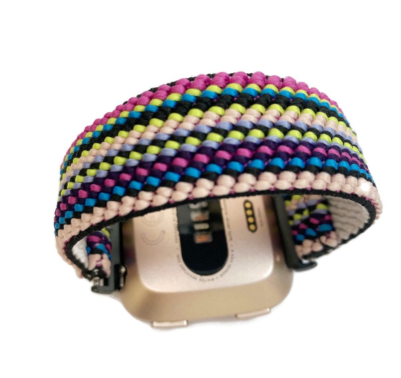 May include: A colourful woven watch band with pink, purple, blue, green, black and white stripes. The band is attached to a gold-coloured smartwatch.