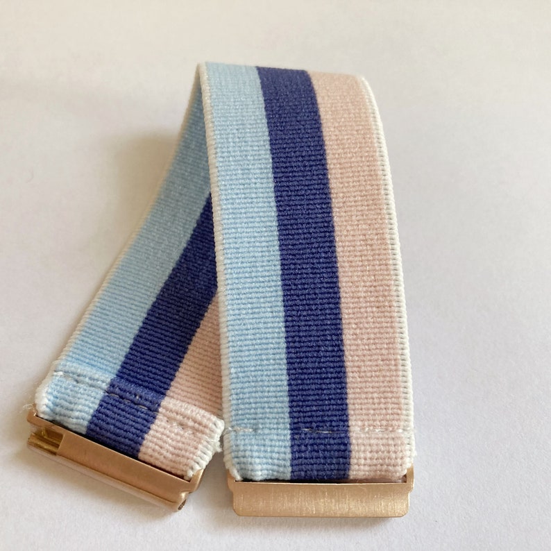 May include: A striped elastic headband with blue, light blue, and pink stripes. The headband has gold metal clasps on each end.