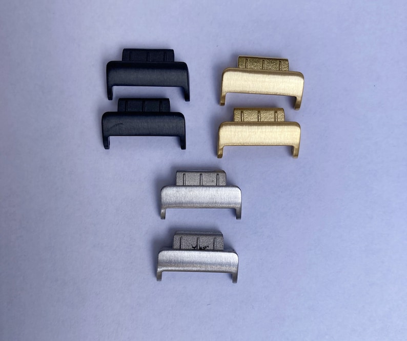 Peut inclure: Divers assortiments de fermoirs de montre noirs, dor&eacute;s et argent&eacute;s. Les fermoirs sont rectangulaires avec une base incurv&eacute;e et un dessus textur&eacute;. Ils sont dispos&eacute;s sur un fond violet clair.