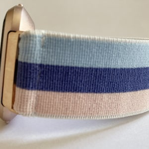 Peut inclure: Une montre connectée rose gold avec un bracelet en tissu rayé rose, bleu et bleu clair.