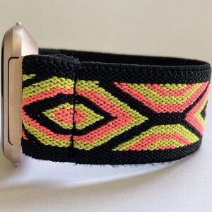 May include: A black elastic watch band with a geometric pattern in pink and yellow. The band is attached to a rose gold watch face.