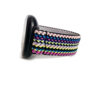 Pode incluir: Um smartwatch preto com uma banda tecida colorida em listras. A banda tem listras rosa, roxas, azuis, verdes e amarelas.