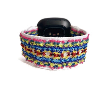 K&ouml;nnte beinhalten: Ein wei&szlig;es Armband mit einem bunten gewebten Muster. Das Armband ist in verschiedenen Farben gehalten, darunter Rot, Gelb, Blau, Gr&uuml;n und Lila. Das Armband ist f&uuml;r eine Smartwatch konzipiert.