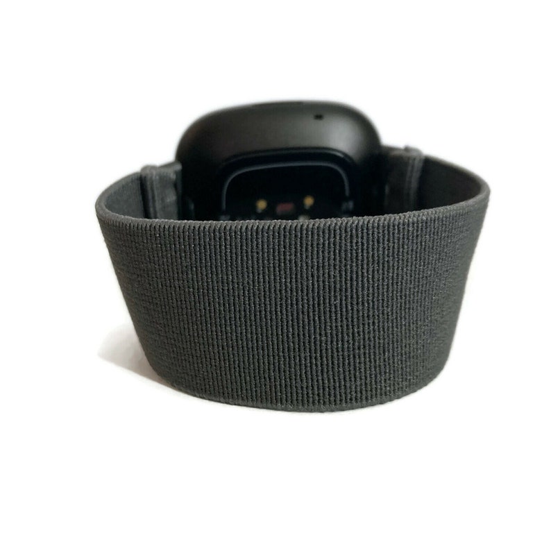Pode incluir: Um rastreador de fitness preto com uma banda el&aacute;stica cinza. O rastreador tem uma pequena tela e um sensor de frequ&ecirc;ncia card&iacute;aca.
