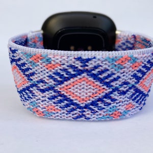 May include: A white, blue, and pink patterned woven watch band. The band is made of a soft, stretchy material and is designed to fit most smartwatches.
