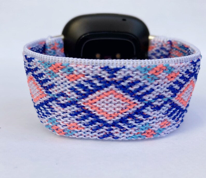 May include: A white, blue, and pink patterned woven watch band. The band is made of a soft, stretchy material and is designed to fit most smartwatches.