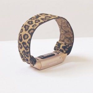 Op de afbeelding: Een goudkleurige fitness tracker met een elastisch bandje met luipaardprint.