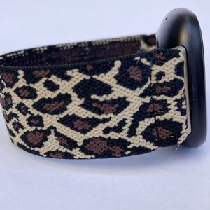 Puede incluir: Una banda de reloj el&aacute;stica de estampado de leopardo negro y marr&oacute;n con una esfera de reloj negra.