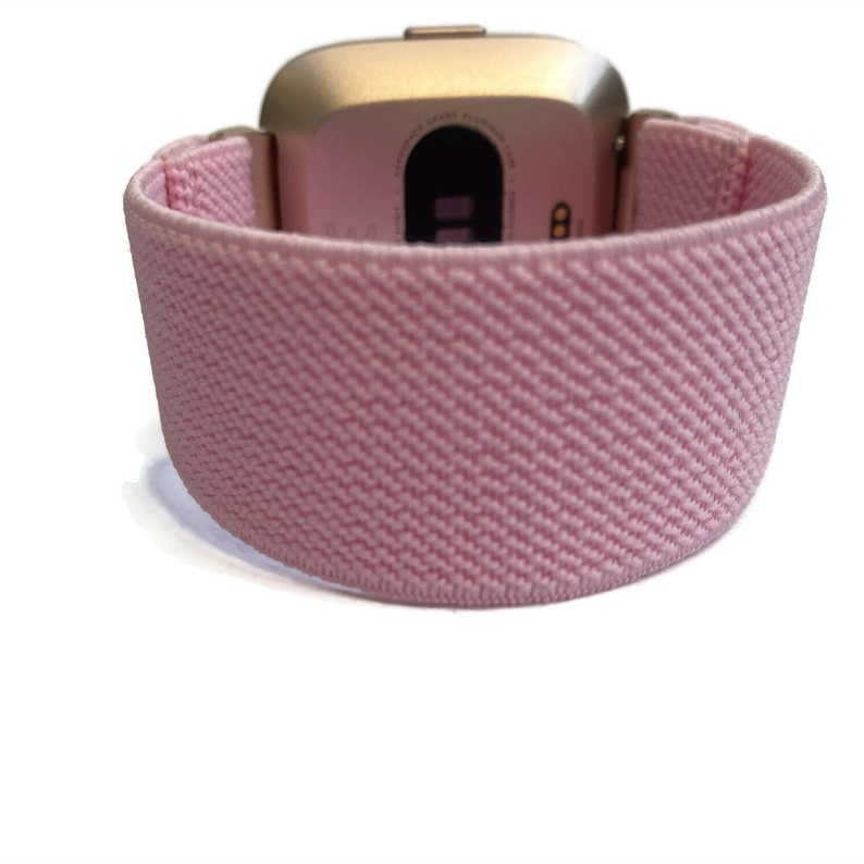May include: A pink elastic watch strap for a rose gold smartwatch. The strap is made of a soft, stretchy material and has a textured surface.
